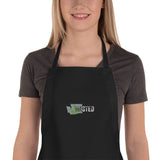 black apron on caucasian female model with gray Washington state outline containing partial cannabis leaf in green, with uppercase "WASTED" across the right of the state, with black WA inside the state outline, and white letters "STED" continuing outside the outline. Center chest placement of embroidered design.
